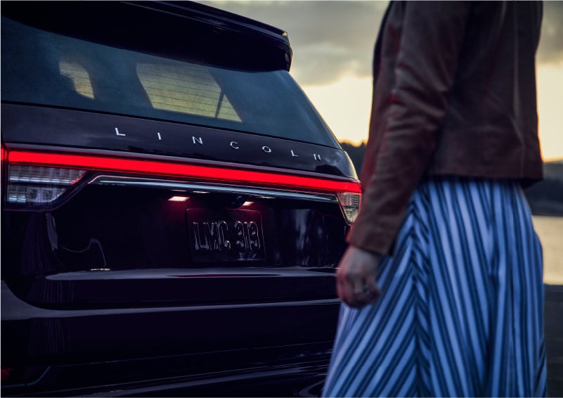 A person is shown near the rear of a 2023 Lincoln Aviator® SUV as the Lincoln Embrace illuminates the rear lights | Gates Lincoln in Richmond KY