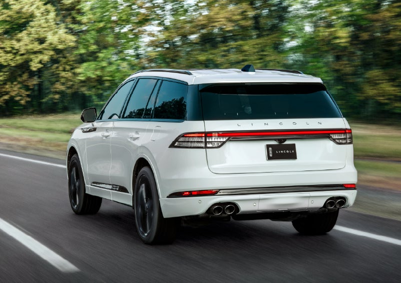 A white 2026 Lincoln Aviator® SUV is being driven away down a road lined with colorful trees. | Gates Lincoln in Richmond KY